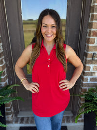 A woman wearing a bright red sleeveless polo shirt with a button placket and blue jeans, standing in front of a brick door with plants visible on either side.