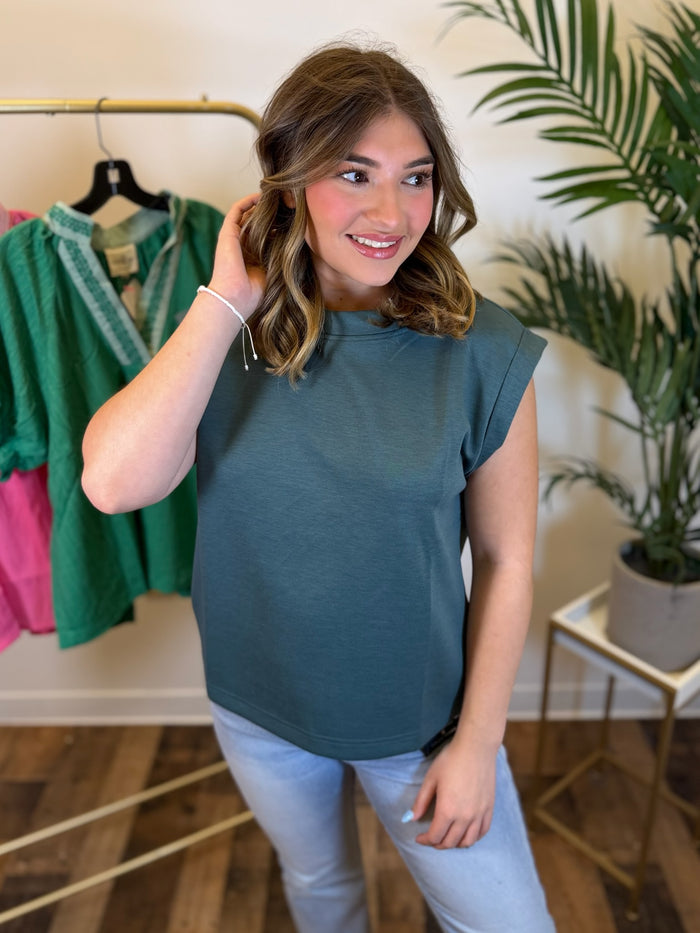 A woman modeling a muted teal sleeveless top with light blue jeans in a boutique setting, with a clothing rack and a potted plant visible in the background.