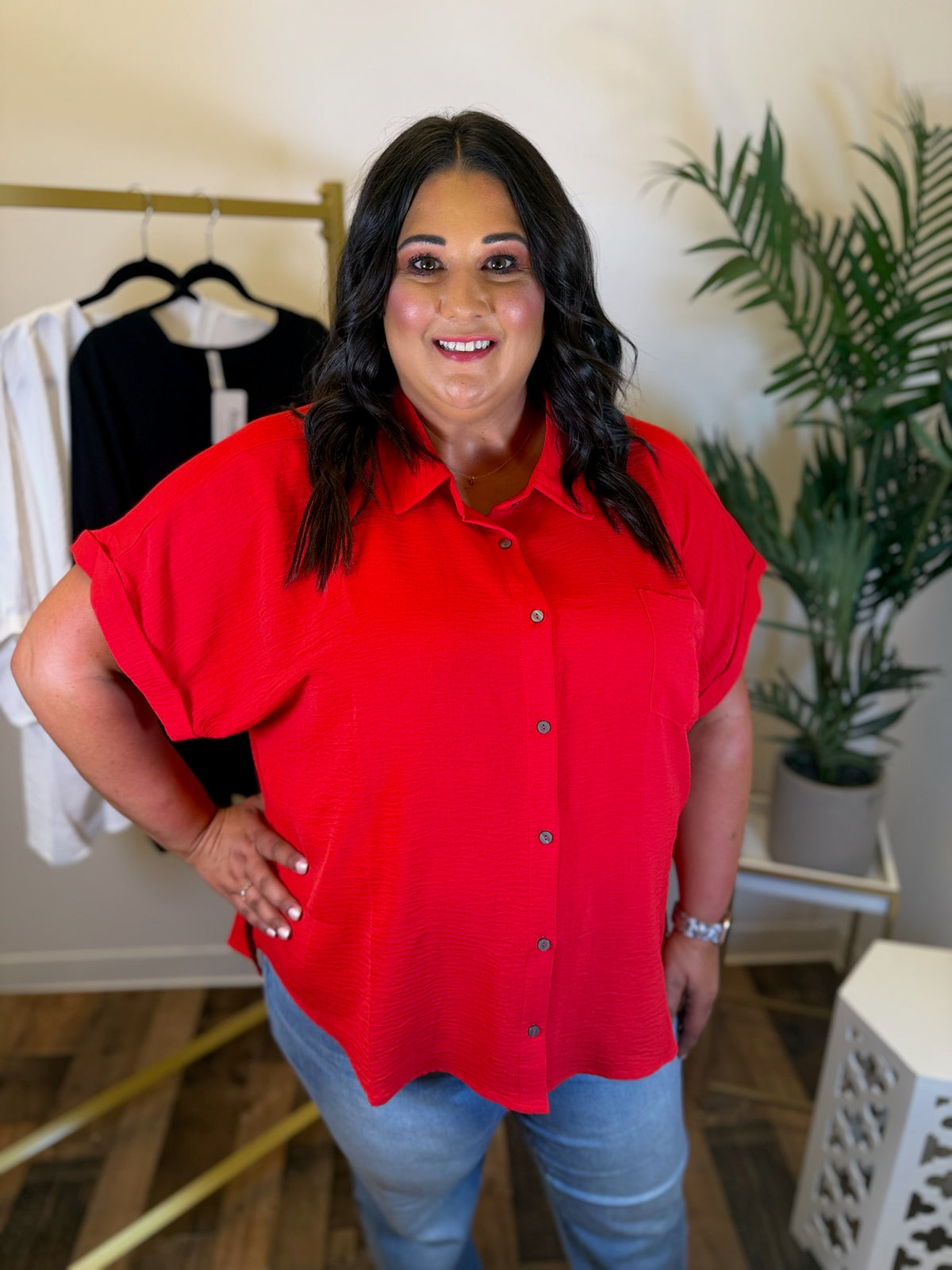 A woman wearing a bright red short-sleeve button-up shirt and light blue jeans, standing in a boutique setting with a clothing rack and a potted plant in the background.