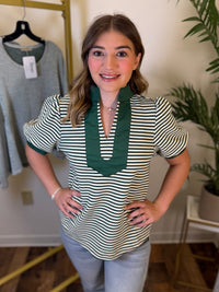 A woman modeling a green and white striped short-sleeve top with a dark green ruffled collar, standing in an indoor boutique setting with a clothing rack and plant in the background.