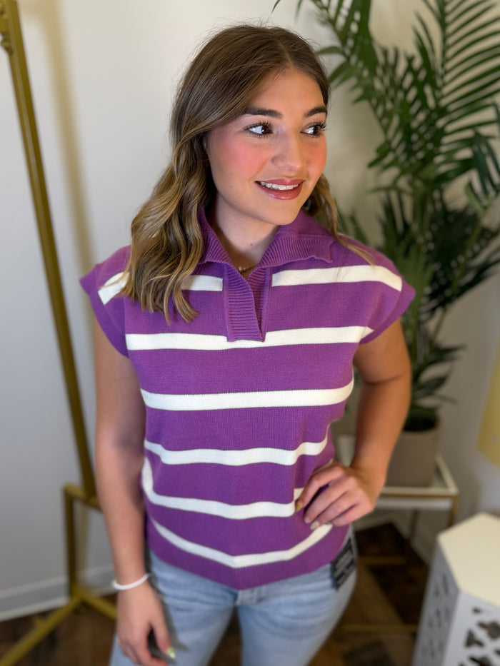 A woman wearing a purple and white horizontally striped short-sleeve knit polo shirt, paired with light blue jeans, standing in an indoor setting with a plant and gold clothing rack visible.