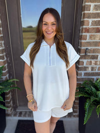 A woman wearing a white short-sleeve zip-front top and matching white shorts, standing outdoors in front of a brick door with plants visible on both sides.