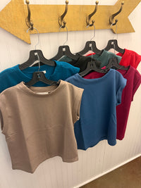 A row of short-sleeve crew-neck crop tops in multiple colors (taupe, teal, blue, magenta, and red) hanging on black hangers from a rustic wooden arrow-shaped rack against a white paneled wall.
