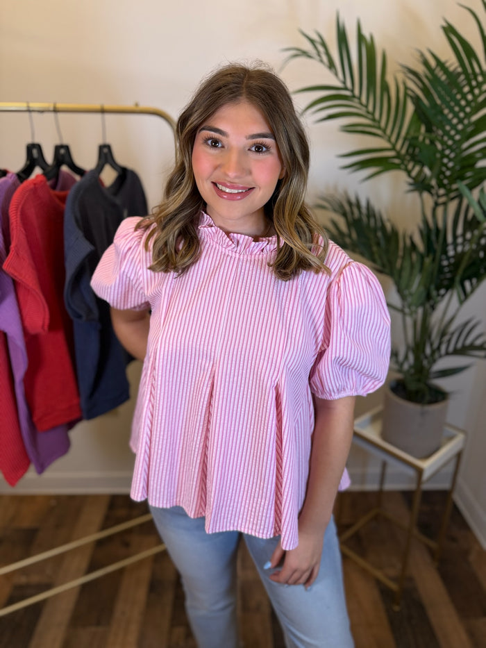 A woman wearing a pink vertically striped puff-sleeve blouse with a ruffled neckline, paired with light blue jeans, standing in a boutique setting with a clothing rack and a potted plant in the background.