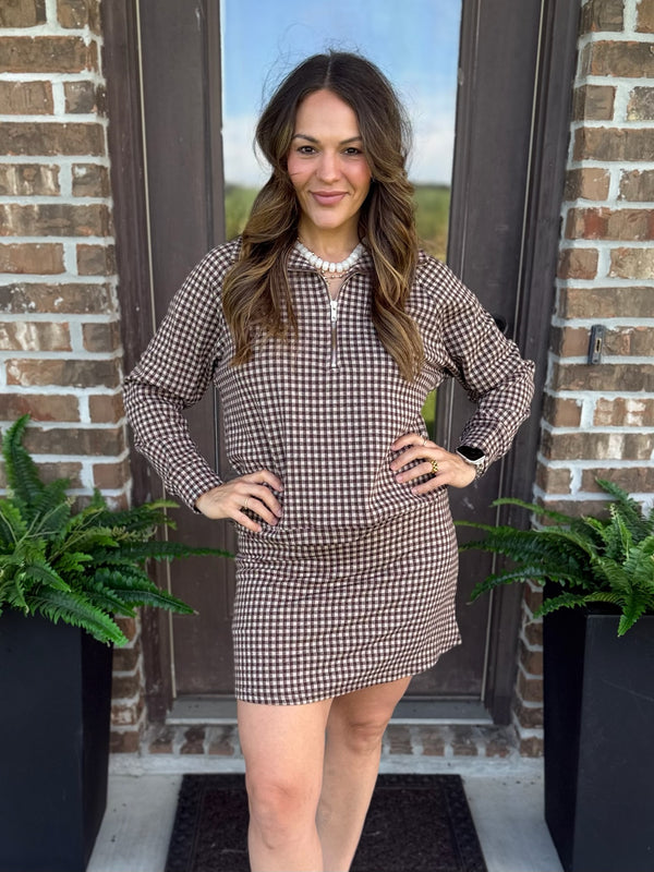 A woman modeling a brown and beige gingham matching skort and shirt set with a half-zip neckline, standing in front of a brick door with potted ferns on either side.