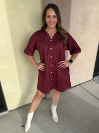 A woman wearing a short-sleeve burgundy button-front shirt dress with scalloped trim and pockets, paired with white ankle boots, standing outdoors against a beige stucco wall.