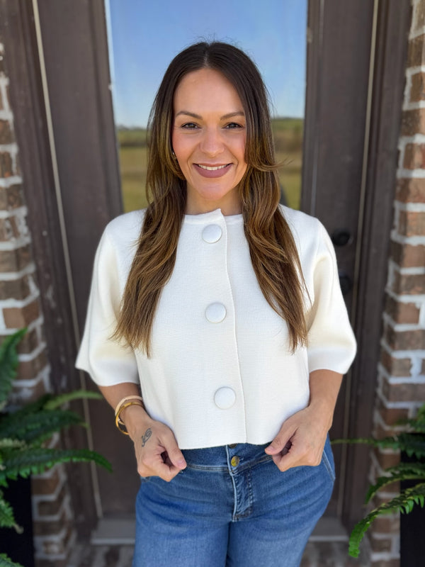 A woman wearing a white short-sleeve cropped cardigan with large decorative buttons and blue jeans, standing in front of a brick doorway.