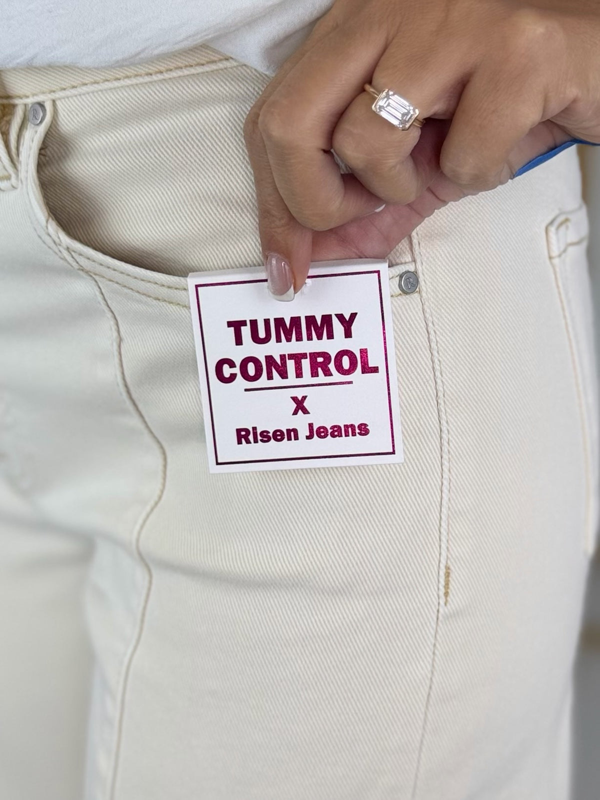 Close-up of a hand holding a 'Tummy Control' jeans tag against beige pants.