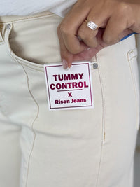 Close-up of a hand holding a 'Tummy Control' jeans tag against beige pants.