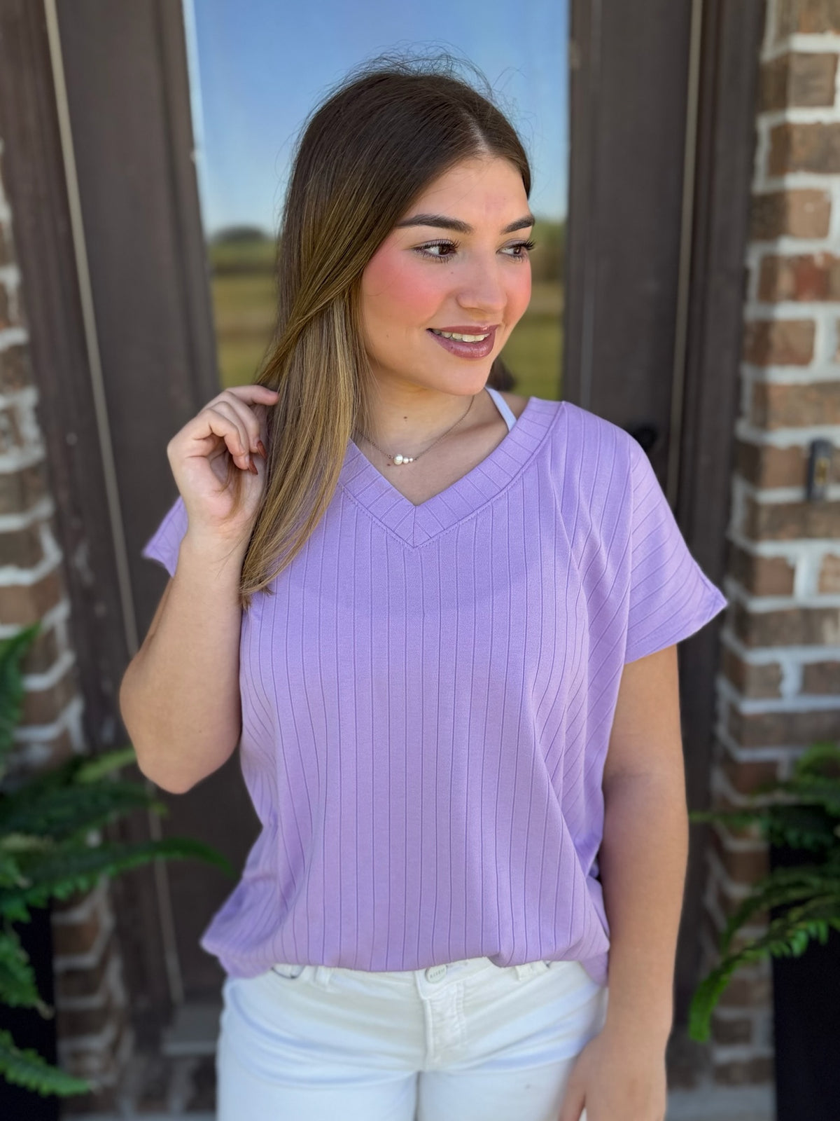 A woman wearing a ribbed lavender V-neck top with short sleeves stands in front of a brick wall.