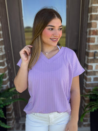 A woman wearing a ribbed lavender V-neck top with short sleeves stands in front of a brick wall.