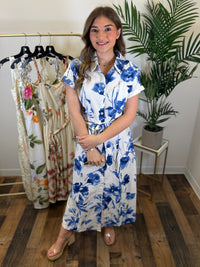 Woman wearing a blue and white floral midi dress with collar neckline standing in a room with a rack of dresses and a plant.