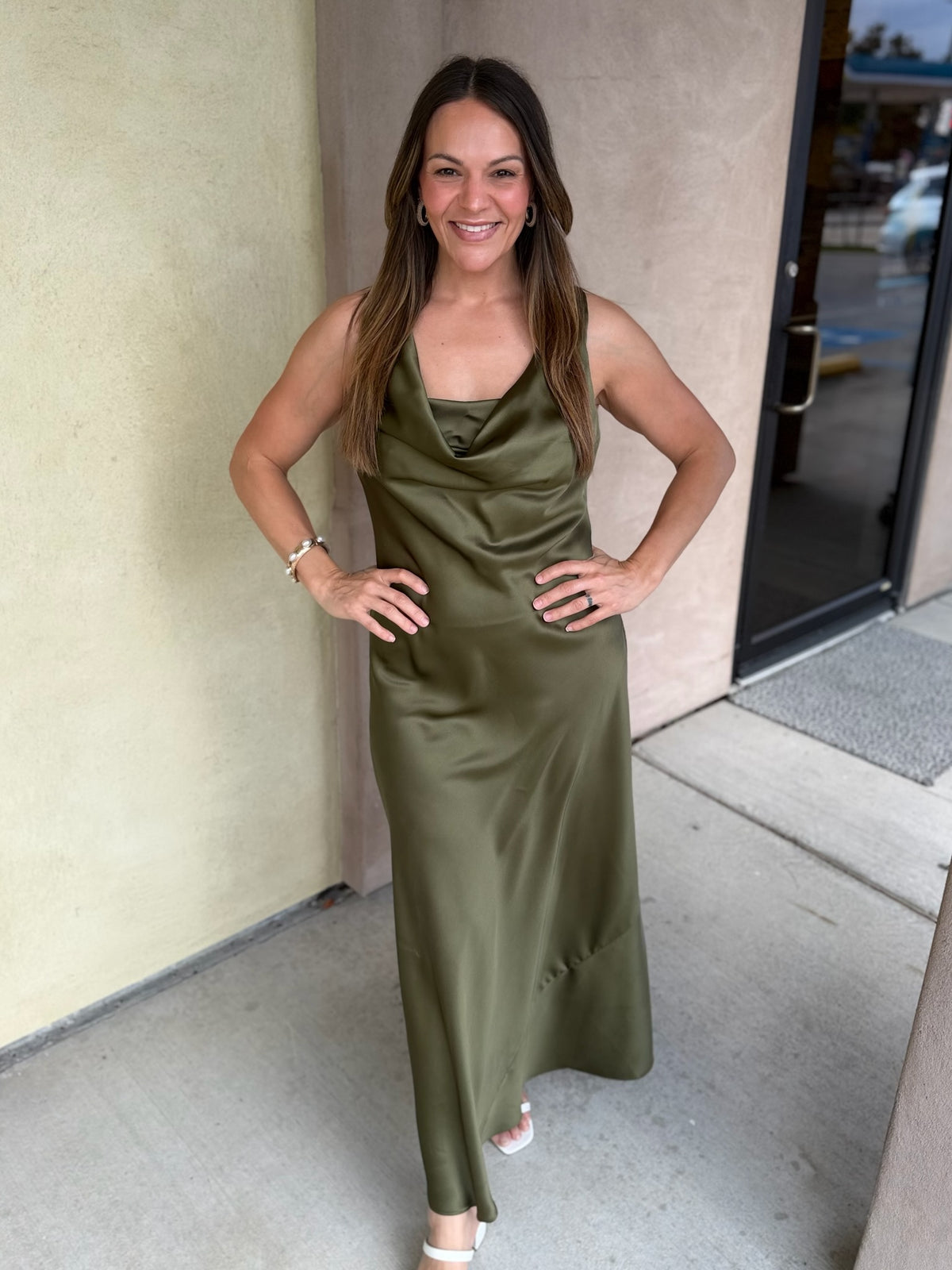 Olive green satin midi dress with spaghetti straps and draped neckline worn by a woman standing in front of beige building.
