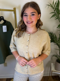 A woman wearing a beige short-sleeve knit cardigan with buttons, standing in a boutique setting with a clothing rack and a potted plant in the background.