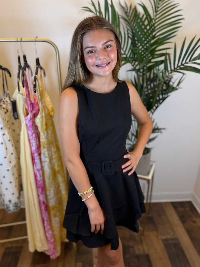 Woman in a short black sleeveless dress with a matching belt standing in a clothing store with racks of dresses and plants in the background.