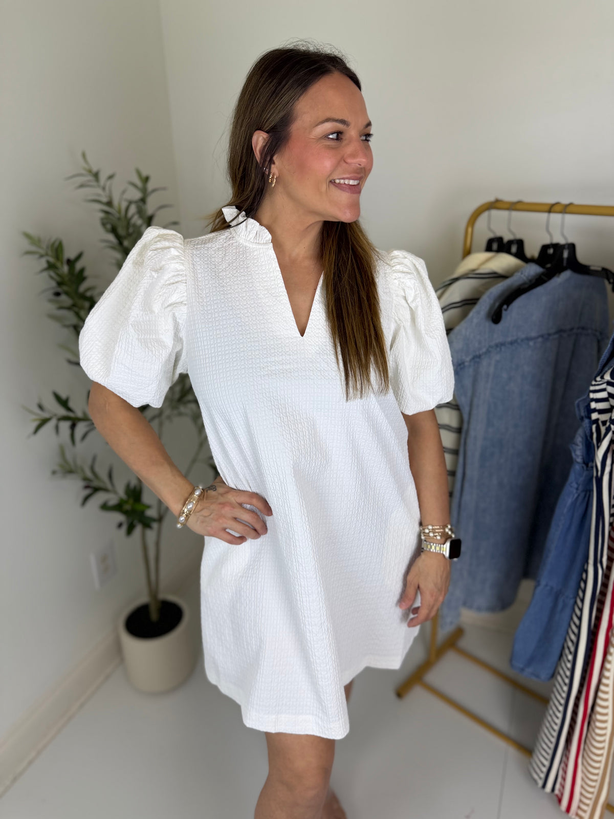 Woman wearing a white textured mini dress with ruffled split neck and voluminous sleeves standing in a room with clothing racks and plants.