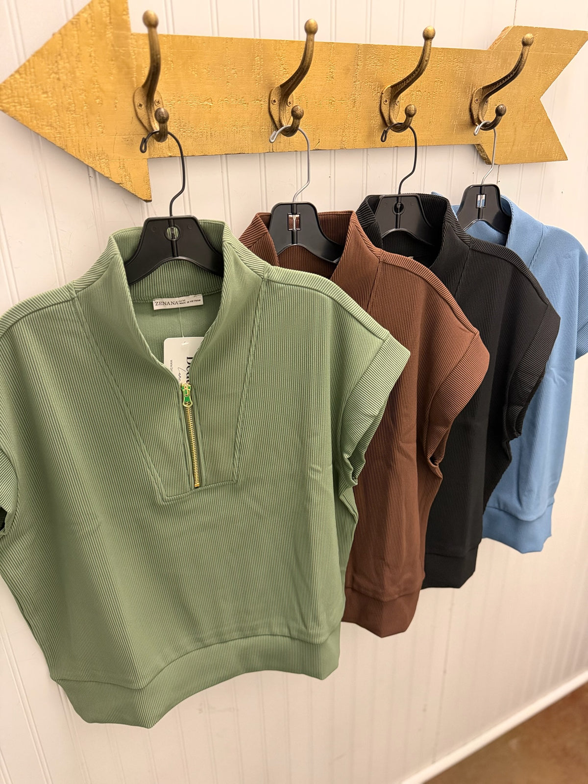 Four women's short-sleeve ribbed pullover tops in sage green, brown, black, and light blue, hanging on black hangers from a gold arrow-shaped wall rack against a white paneled wall.