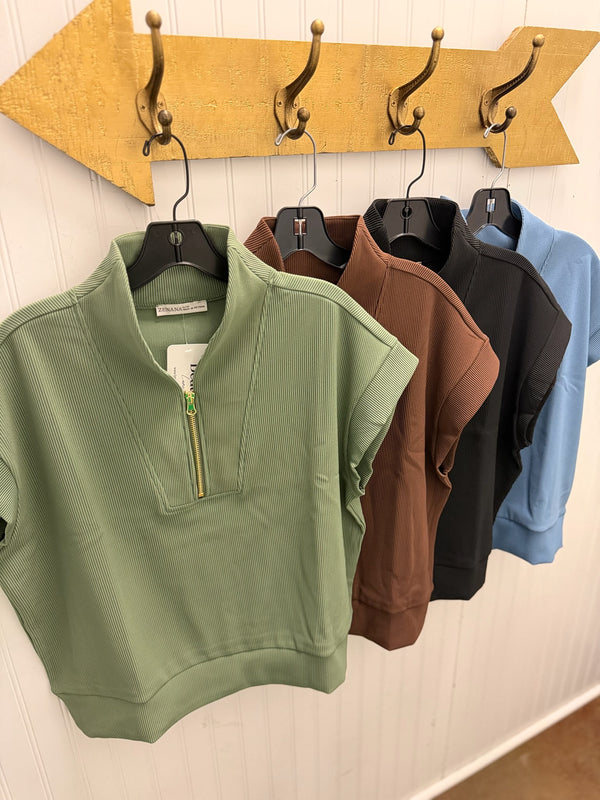 Four women's short-sleeve ribbed pullover tops in sage green, brown, black, and light blue, hanging on black hangers from a gold arrow-shaped wall rack against a white paneled wall.