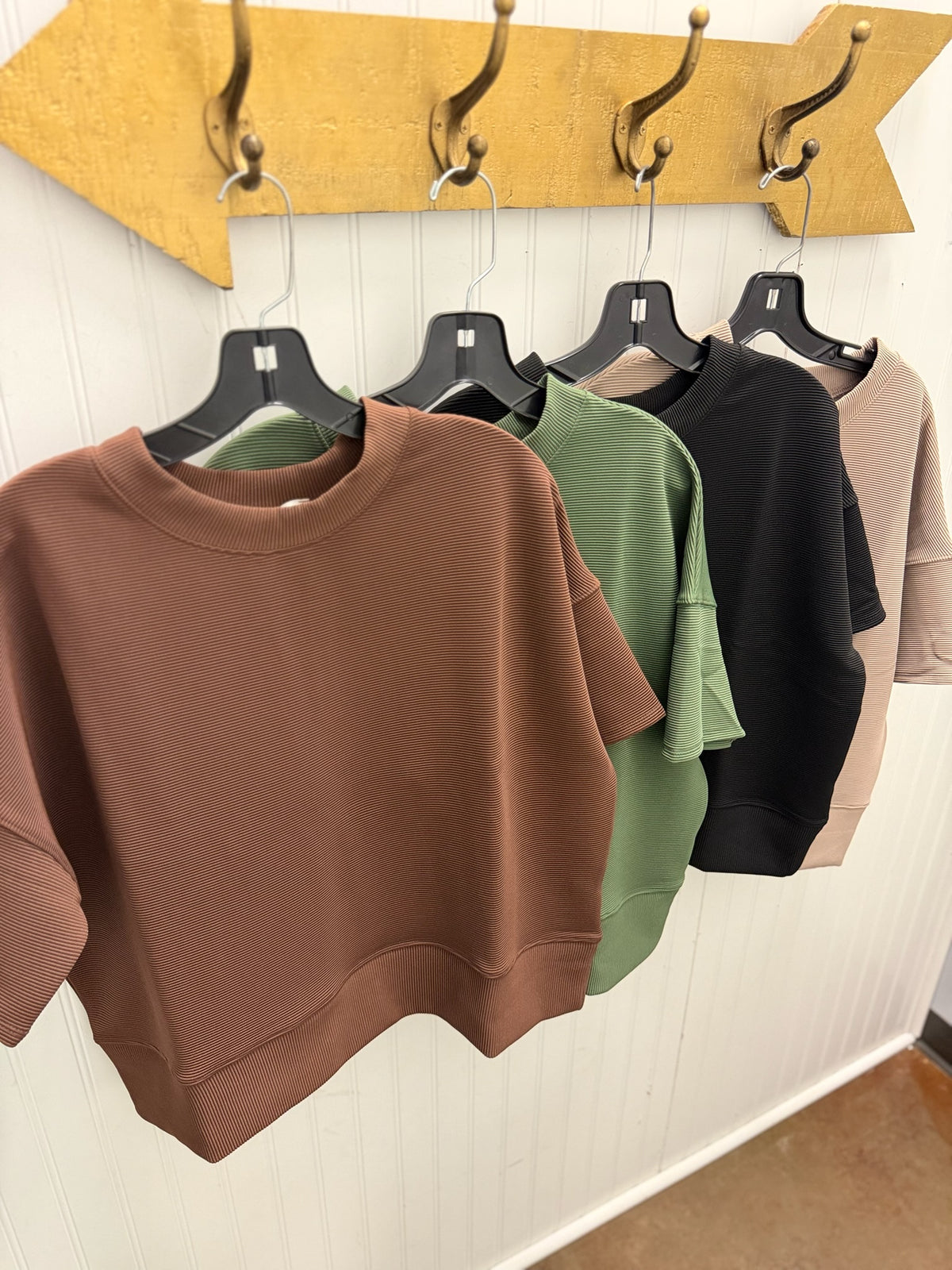 Four cropped crewneck sweatshirts in brown, sage green, black, and beige hang on black hangers from a gold arrow-shaped wall rack against a white paneled wall.