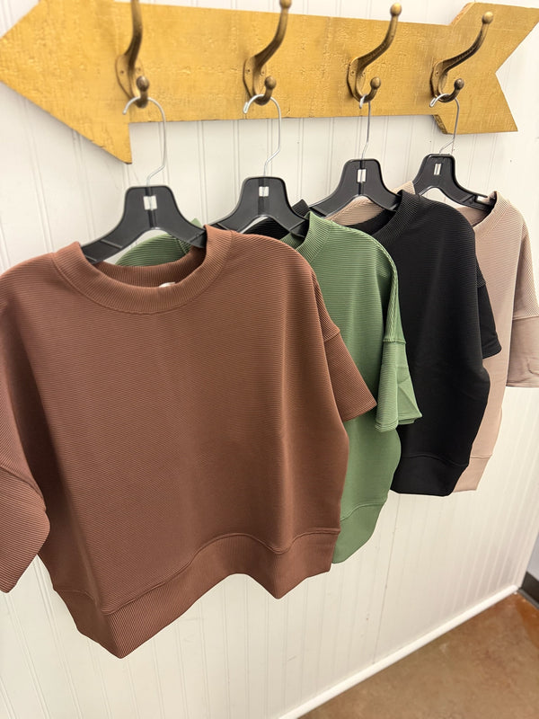 Four cropped crewneck sweatshirts in brown, sage green, black, and beige hang on black hangers from a gold arrow-shaped wall rack against a white paneled wall.