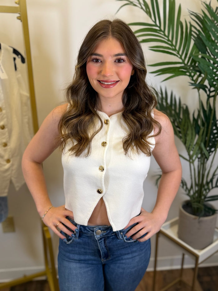 Woman wearing a white blouse with gold buttons and blue jeans in a room with plants.