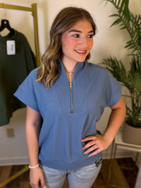 A woman wearing a dusty blue short-sleeve quarter-zip top with a stand collar, paired with light-wash jeans, standing in an indoor boutique setting.