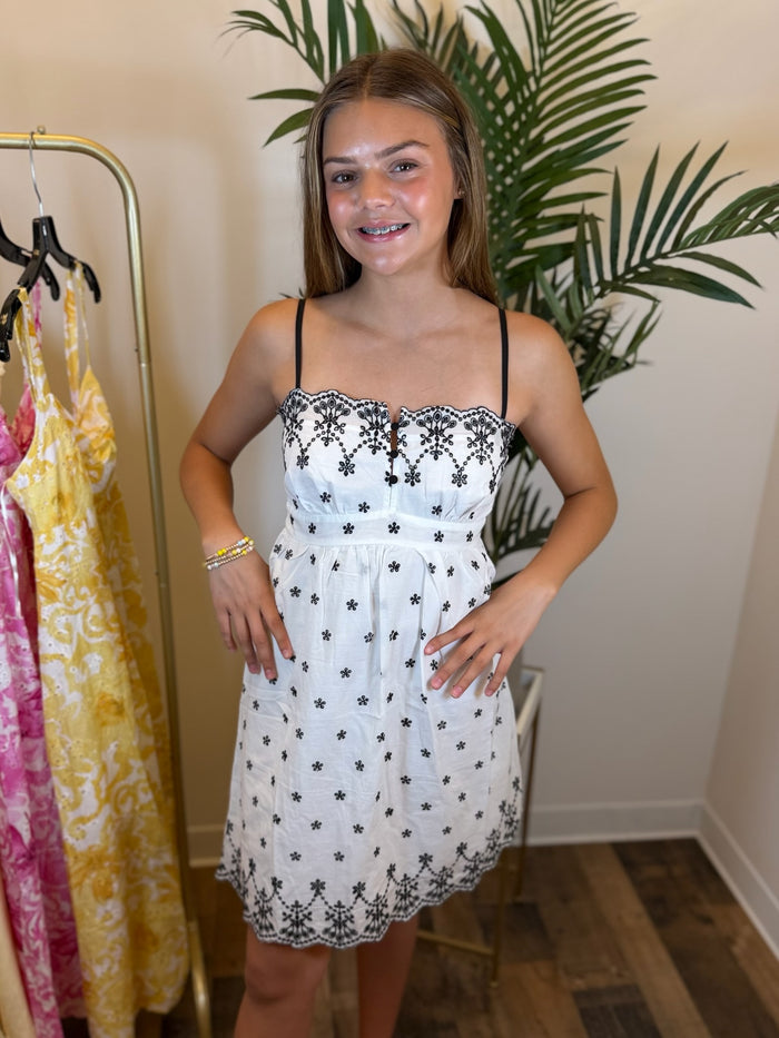 Young girl wearing a short white dress with black floral patterns and spaghetti straps in a room with clothes and plants.