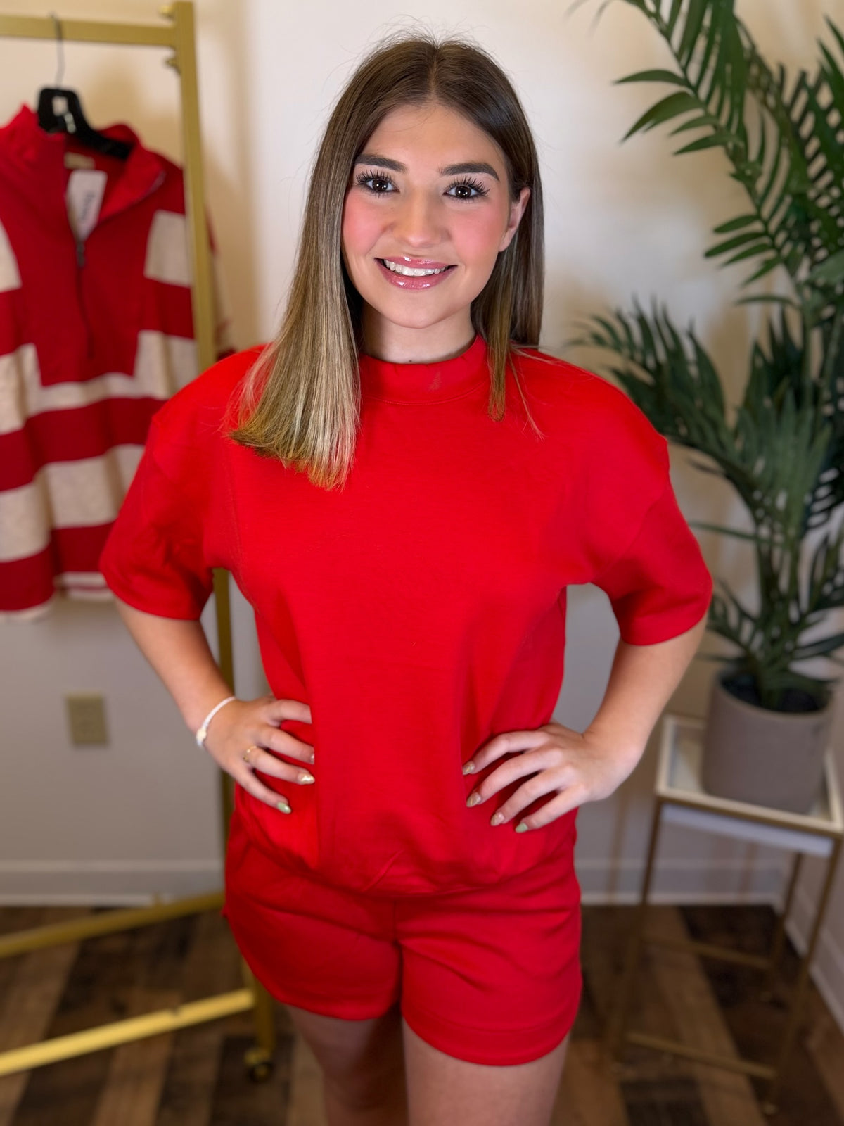A woman modeling a bright red short-sleeve mock neck top and matching red shorts in an indoor setting with a clothing rack and a potted plant in the background.