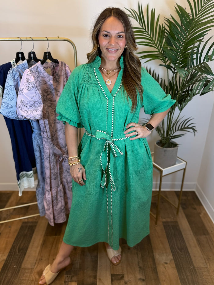 Woman in a textured green midi dress with split neckline, puff sleeves, and adjustable waist tie standing in a room with clothing racks and plants.