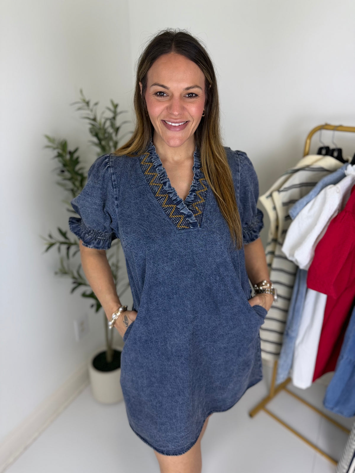 Woman wearing a blue dress with decorative v  neckline in a room with a plant and clothing rack.