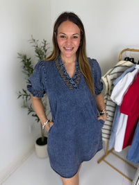 Woman wearing a blue dress with decorative v  neckline in a room with a plant and clothing rack.