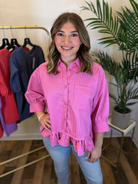 A woman modeling a pink and white vertically striped button-down shirt with puff sleeves and a ruffled hem, paired with light blue jeans in a boutique setting.