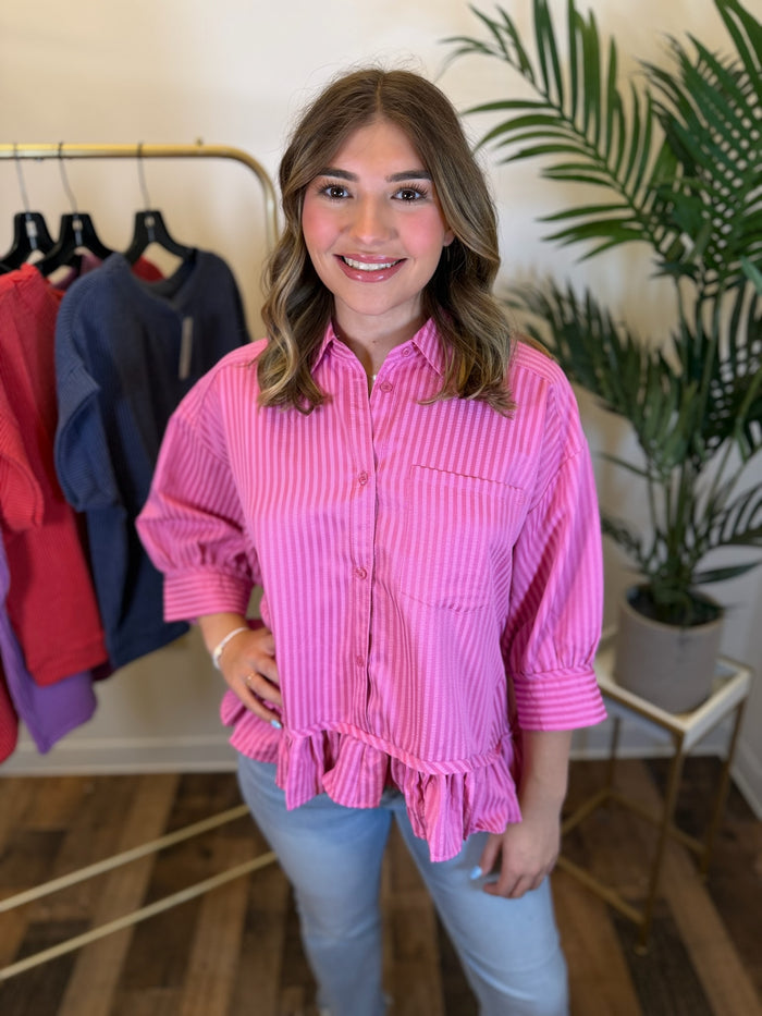A woman modeling a pink and white vertically striped button-down shirt with puff sleeves and a ruffled hem, paired with light blue jeans in a boutique setting.