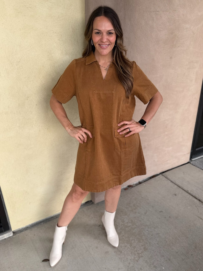A woman wearing a short-sleeve camel utility shirt dress with white ankle boots, posing outdoors against a beige stucco wall.