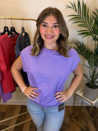 A woman wearing a lavender purple sleeveless t-shirt with raw trimmed neckline and light blue jeans, standing in a boutique setting with a clothing rack and a potted plant in the background.