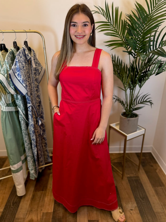 Woman in a red sleeveless maxi dress  with white stitch trim and square neckline standing in a clothing store with plants and racks of clothes in the background.