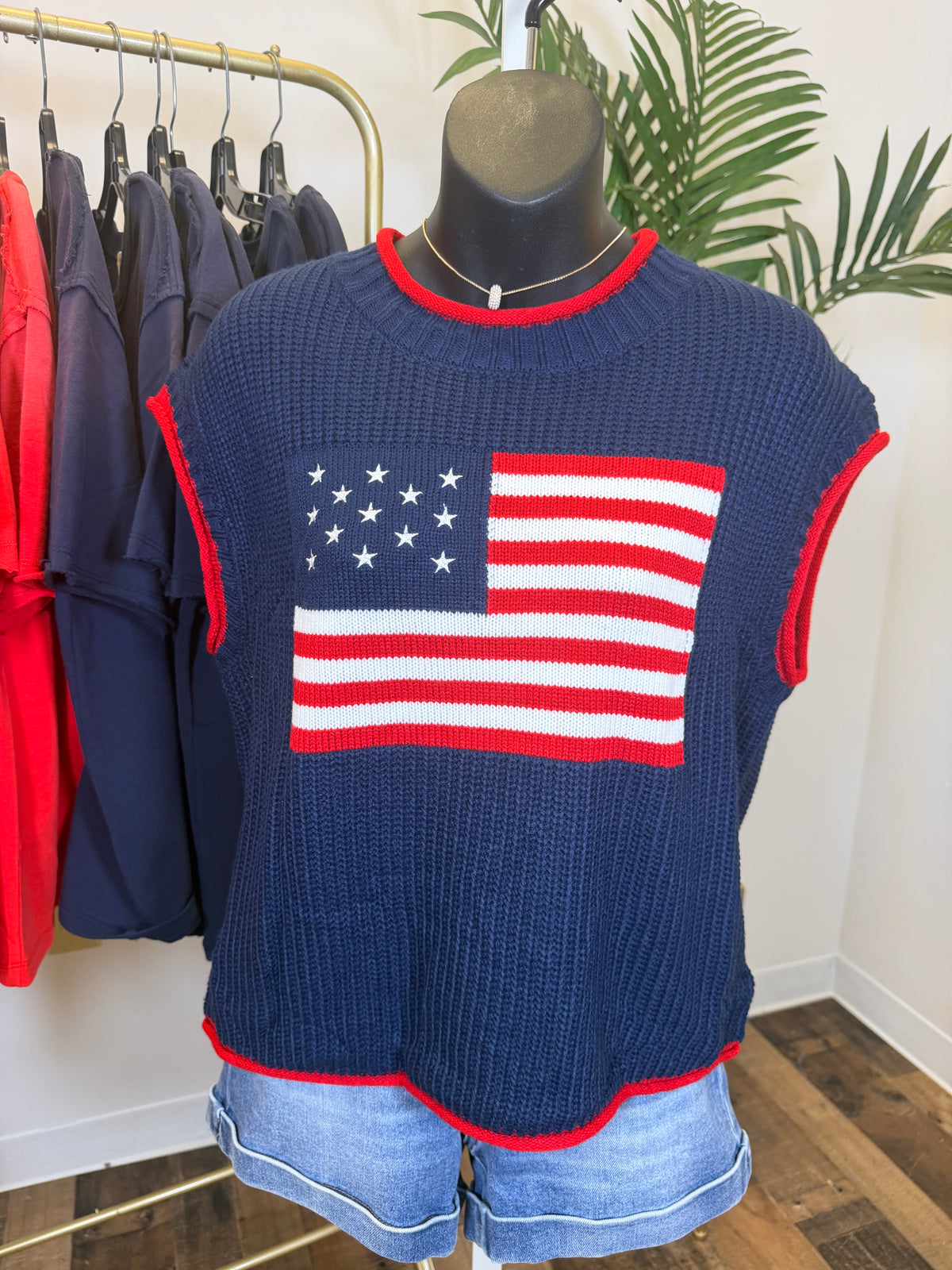 Mannequin wearing a navy sweater with an American flag design and denim shorts, surrounded by hanging clothes and shoes.