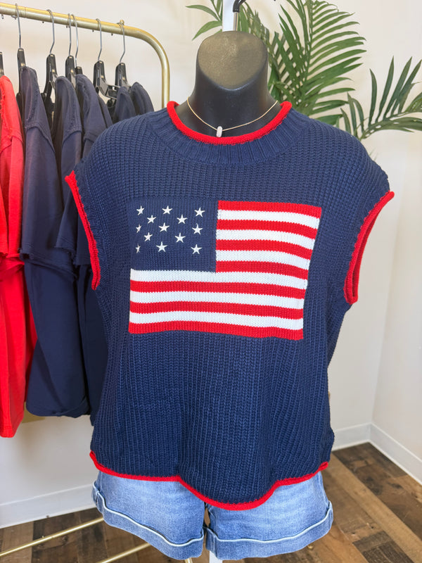 Mannequin wearing a navy sweater with an American flag design and denim shorts, surrounded by hanging clothes and shoes.