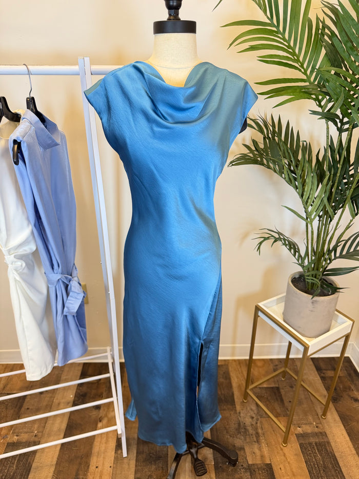Silky Blue jewel-toned midi dress with short sleeves and draped neckline on a mannequin in front of plants and clothes hanging on rack.