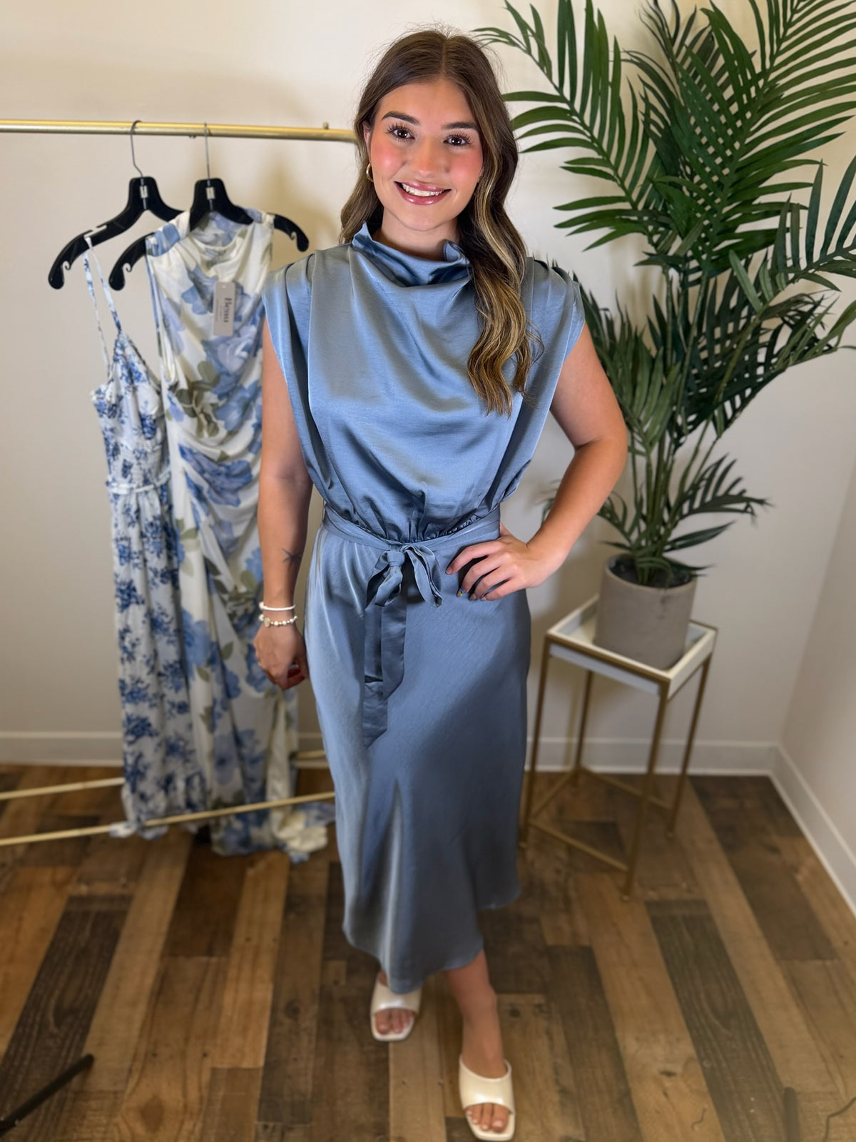 Woman in a slate blue midi dress with draped neckline and adjustable waist tie standing in a room with a rack of dresses and a plant.