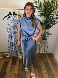 Woman in a slate blue midi dress with draped neckline and adjustable waist tie standing in a room with a rack of dresses and a plant.