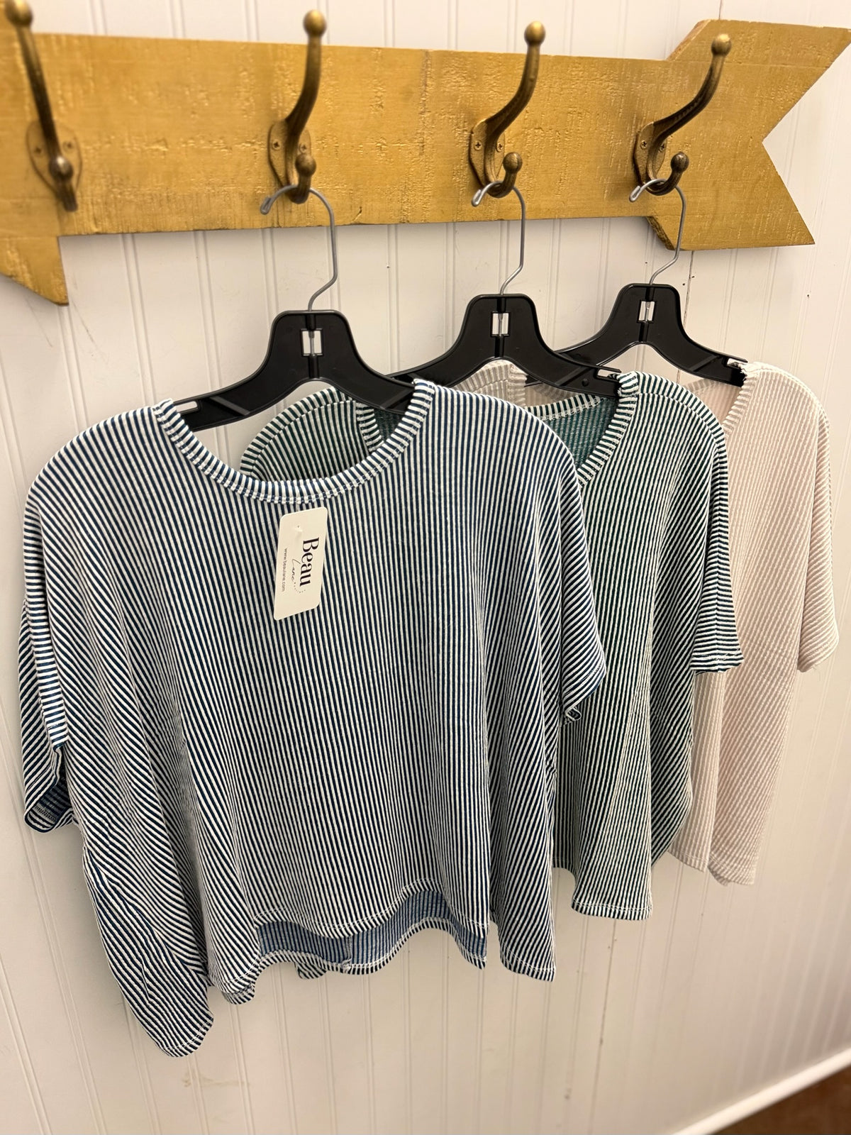 Three women's short-sleeve knit tops hanging on black hangers against a white paneled wall, with a wooden coat rack above. The front top is blue and white vertically striped, the middle top is a muted sage green, and the back top is a light beige/cream.