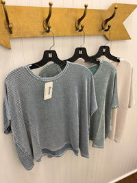 Three women's short-sleeve knit tops hanging on black hangers against a white paneled wall, with a wooden coat rack above. The front top is blue and white vertically striped, the middle top is a muted sage green, and the back top is a light beige/cream.