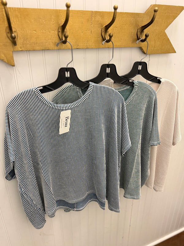 Three women's short-sleeve knit tops hanging on black hangers against a white paneled wall, with a wooden coat rack above. The front top is blue and white vertically striped, the middle top is a muted sage green, and the back top is a light beige/cream.