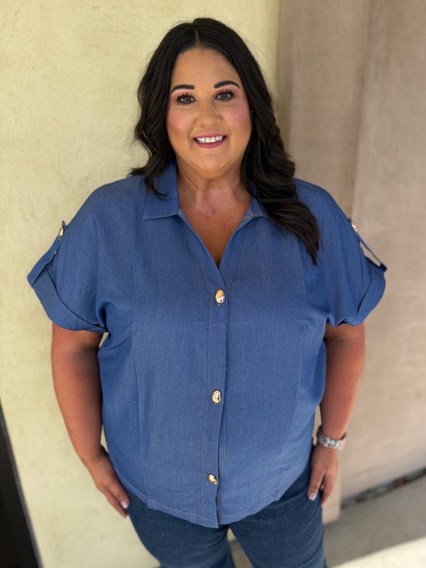 A woman wearing a blue short-sleeve button-up shirt with gold buttons and blue jeans, standing against a beige wall.