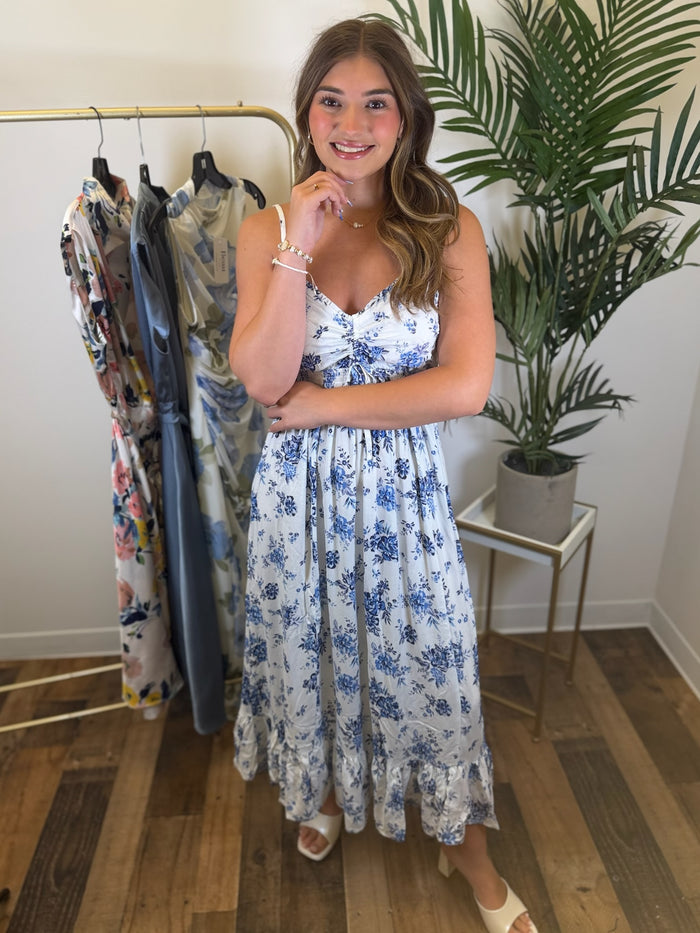 Woman in a sleeveless white maxi dress  with blue floral print and sweetheart neckline standing in a room with clothing racks and plants.