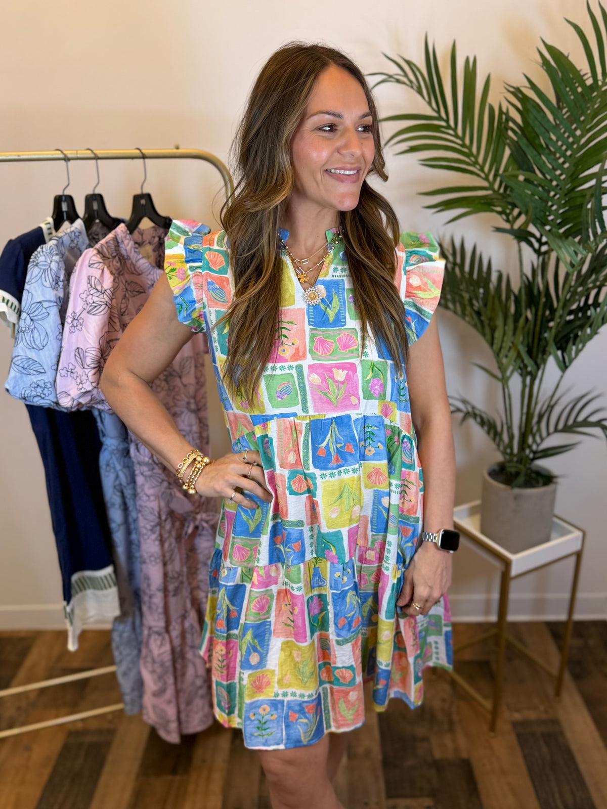 Woman wearing a colorful patterned dress standing in a clothing store with racks and plants in the background.