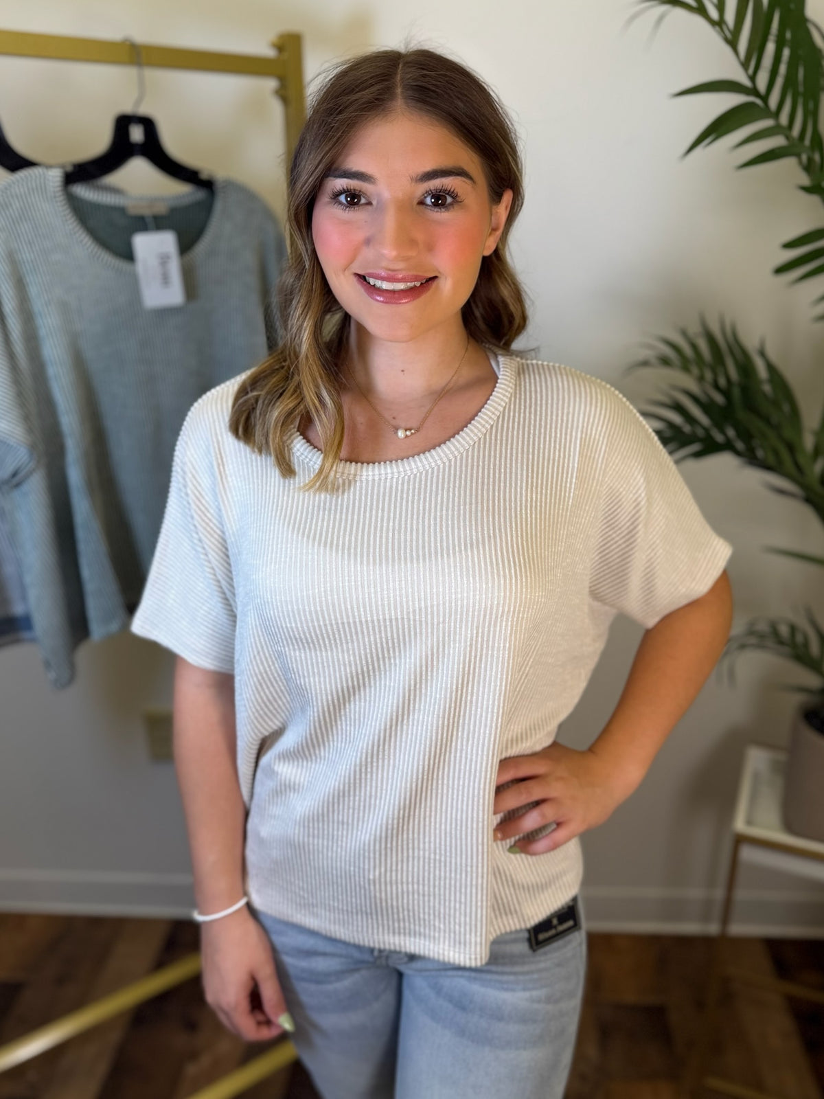 A woman wearing a cream-colored ribbed short-sleeve t-shirt and light blue jeans, standing in a room with a clothing rack and a plant in the background.