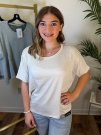 A woman wearing a cream-colored ribbed short-sleeve t-shirt and light blue jeans, standing in a room with a clothing rack and a plant in the background.