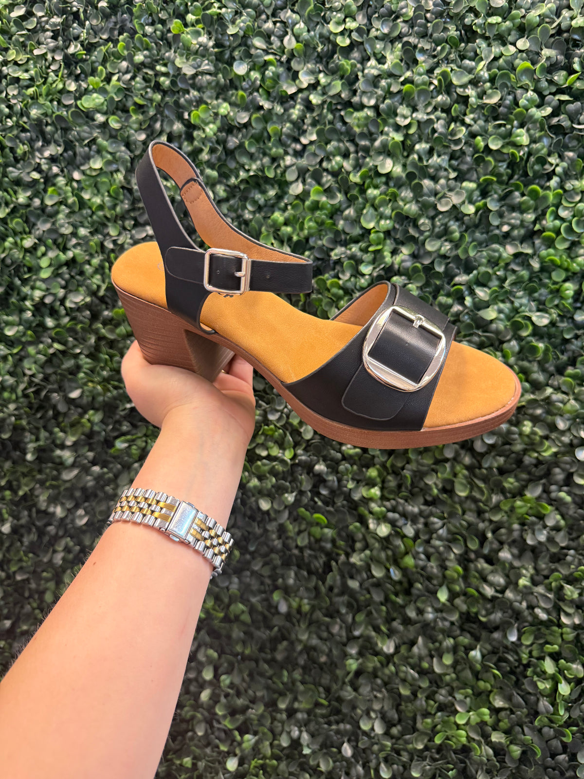 Hand holding a sandal with a buckle design against a green textured background